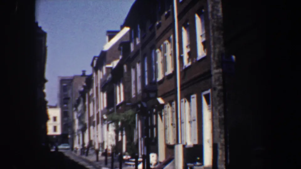 1962 Philadelphia home-movie frame of a narrow row-house street with brick facades, shutters, street lamps, and tiny distant figures or objects near the sidewalk.