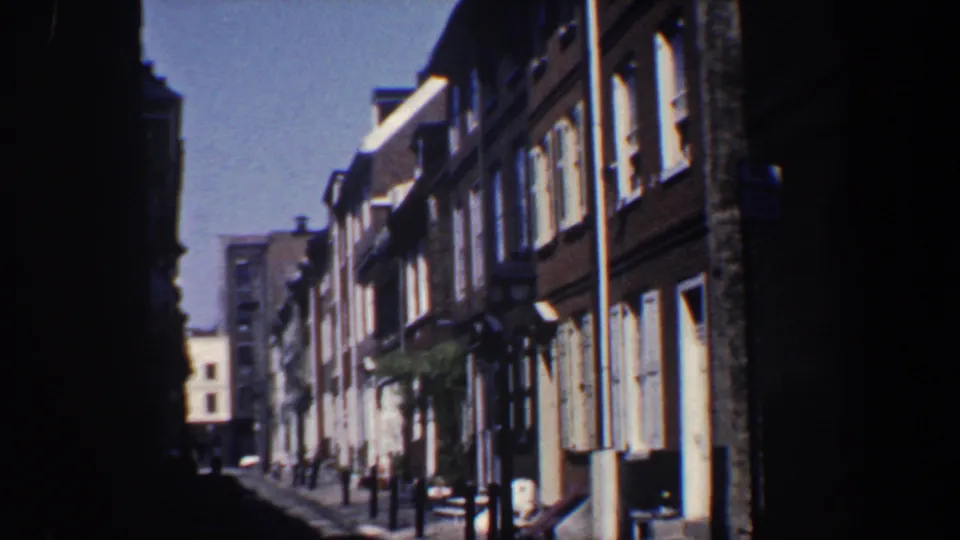 1962 Philadelphia frame looking along a compact row-house block, with brick fronts, white shutters, street lamps, and strong shadow on both sides of the image.