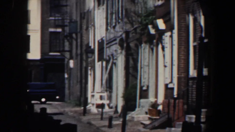 1962 Philadelphia frame looking down a narrow residential street with brick buildings, fire escape, streetlamp, and a dark van.