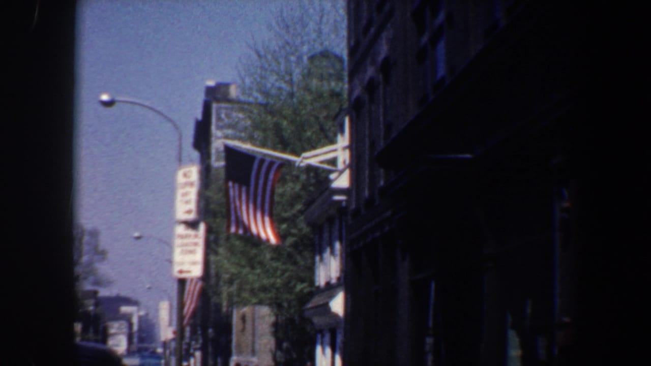 A 13-star American flag hangs from a Philadelphia building facade in 1962, with street signs, trees, and older cars visible beyond a dark foreground edge.