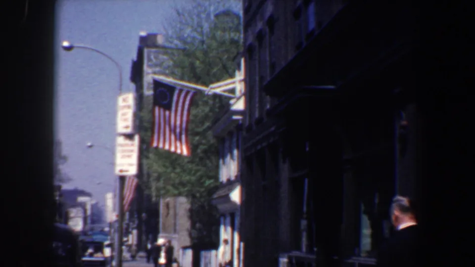 A 13-star American flag waves from a Philadelphia building in 1962 as a suited adult man appears near the right edge of the shaded sidewalk view.
