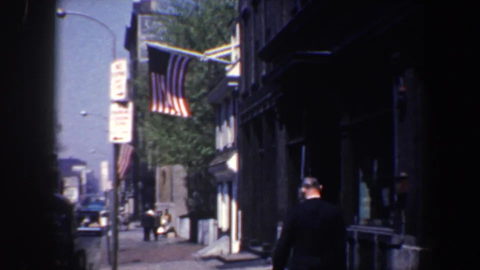 A 1962 Philadelphia street view shows a 13-star American flag above the sidewalk while a suited adult man walks past storefronts and parked or passing cars.