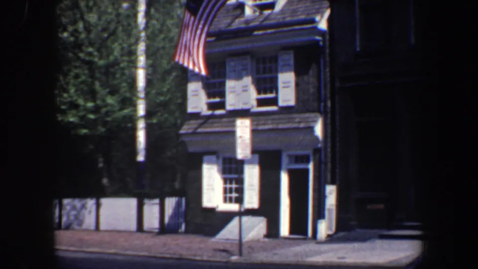 A corner building in 1962 Philadelphia carries a 13-star American flag, with white street signs, windows, a doorway, trees, and deep shadow framing the view.