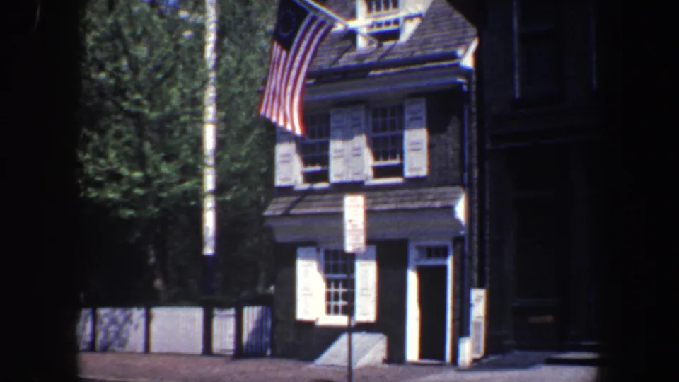 1962 Philadelphia home-movie frame showing a brick colonial-style facade with white shutters, trees, and a Betsy Ross-style flag.