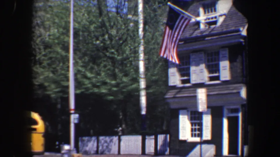 Philadelphia 1962 street frame with a brick building, white shutters, an American flag, trees, and a yellow bus passing at the edge of view.