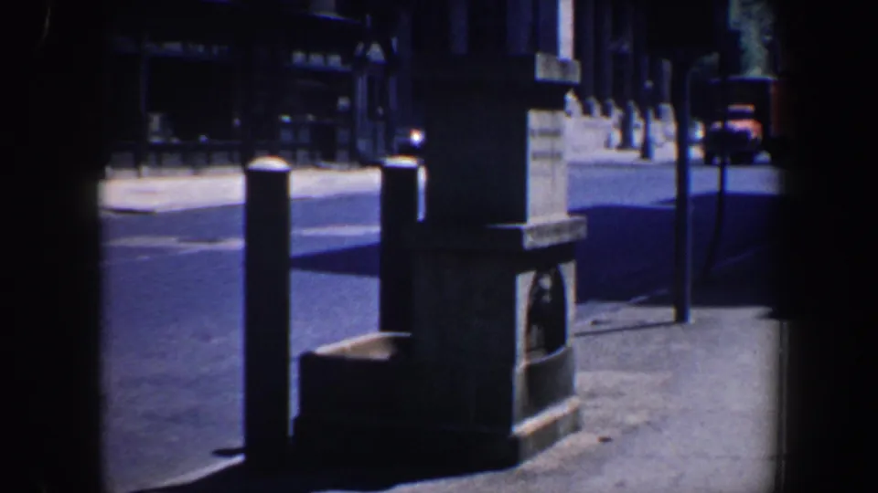 Philadelphia 1962 home-movie view looking past a stone monument and sidewalk posts toward older vehicles on a quiet urban street.