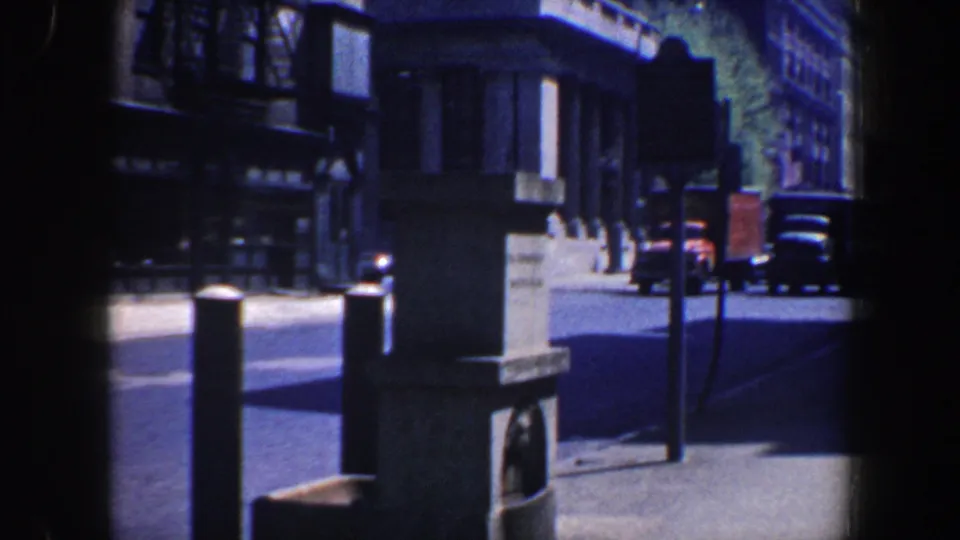 Philadelphia 1962 frame showing a stone monument, bollards, older parked vehicles, and a shaded urban street beside formal building facades.