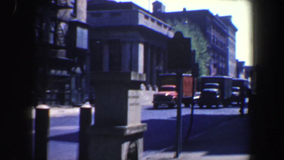 1962 Philadelphia home-movie frame showing classic cars and trucks moving along a sunlit urban street near a columned corner building.