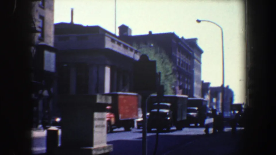 1962 Philadelphia street view with dark 8mm frame borders, a tall streetlamp, delivery trucks, and classic cars passing a corner building.