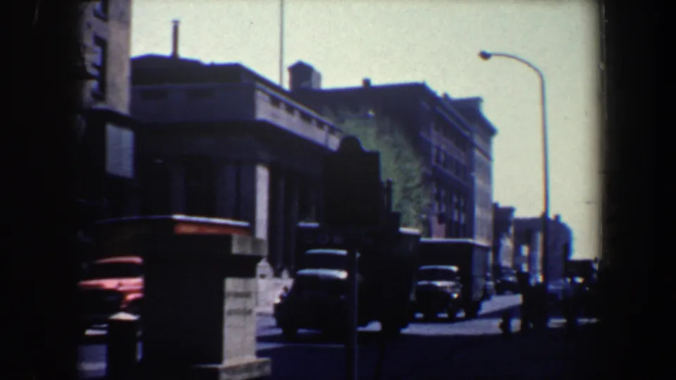 1962 Philadelphia home-movie frame with a red delivery truck, classic cars, street signage, and multi-story urban buildings under bright sky.