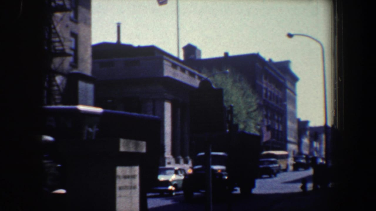 1962 Philadelphia street view on 8mm film, showing brick buildings, parked cars, a streetlamp, and a flag in soft exposure.