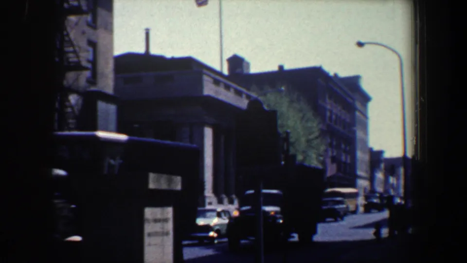 1962 Philadelphia street view on 8mm film, showing brick buildings, parked cars, a streetlamp, and a flag in soft exposure.