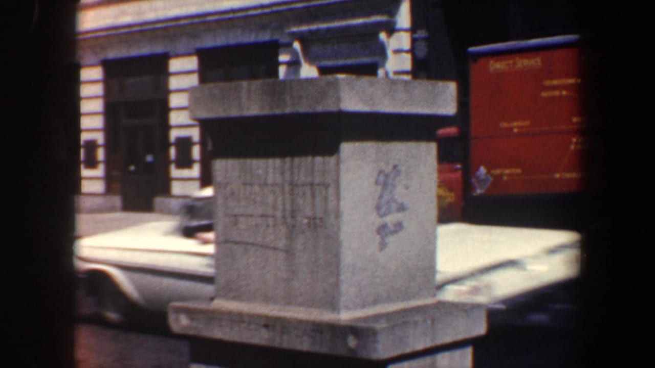 Stone monument beside a city street with parked cars and a building facade behind it.