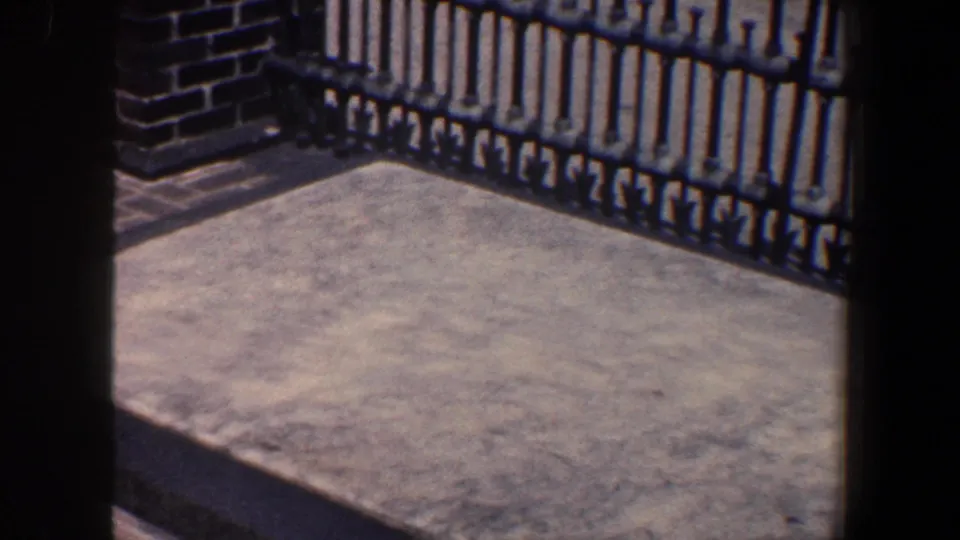 1962 Philadelphia home-movie frame showing a concrete platform bordered by a black iron gate and brickwork.