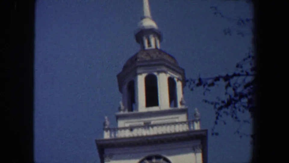 1962 Philadelphia frame showing the cupola, spire, white railing, and upper tower details against a blue sky with branches nearby.