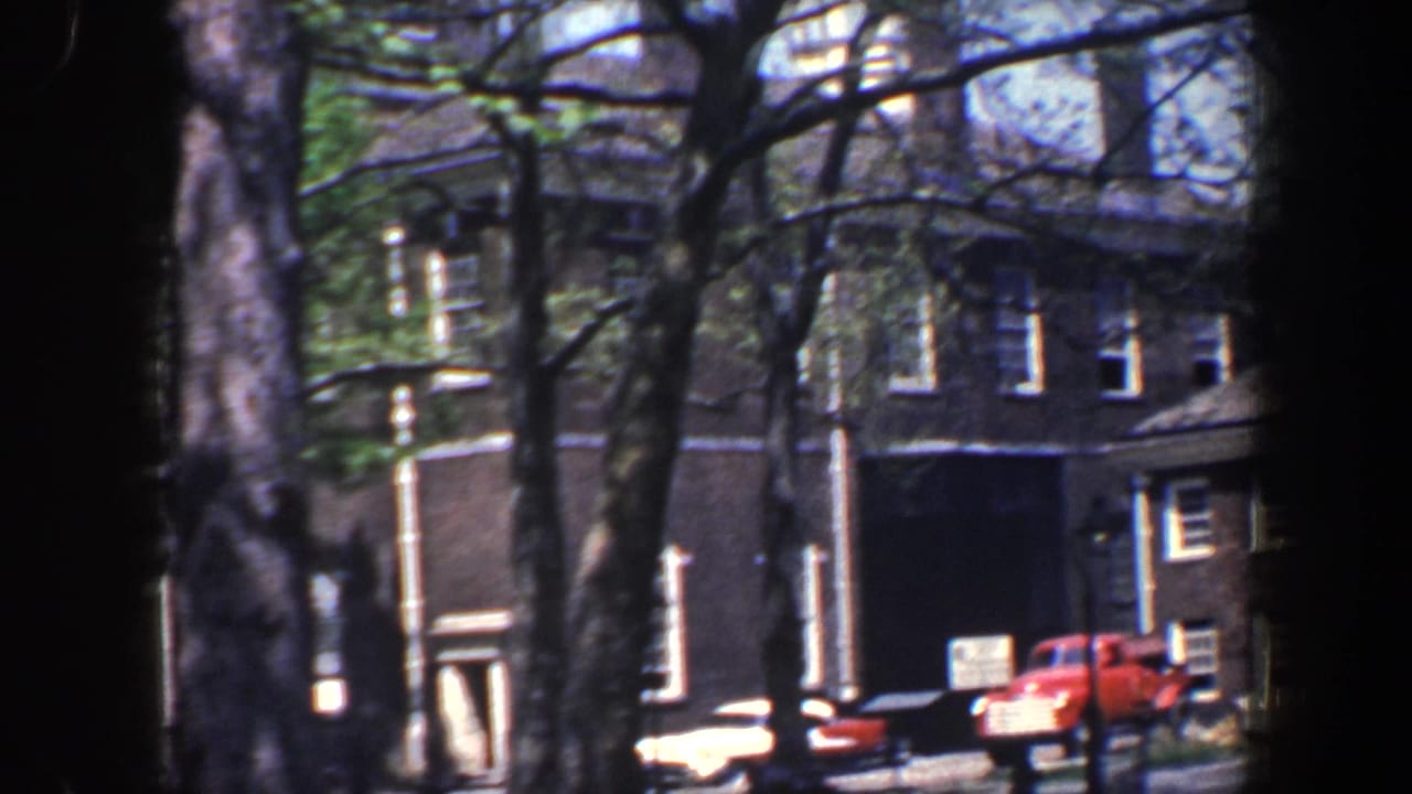 1962 Philadelphia street view with brick facades, parked cars, a red truck, and tree trunks partly blocking the 8mm frame.