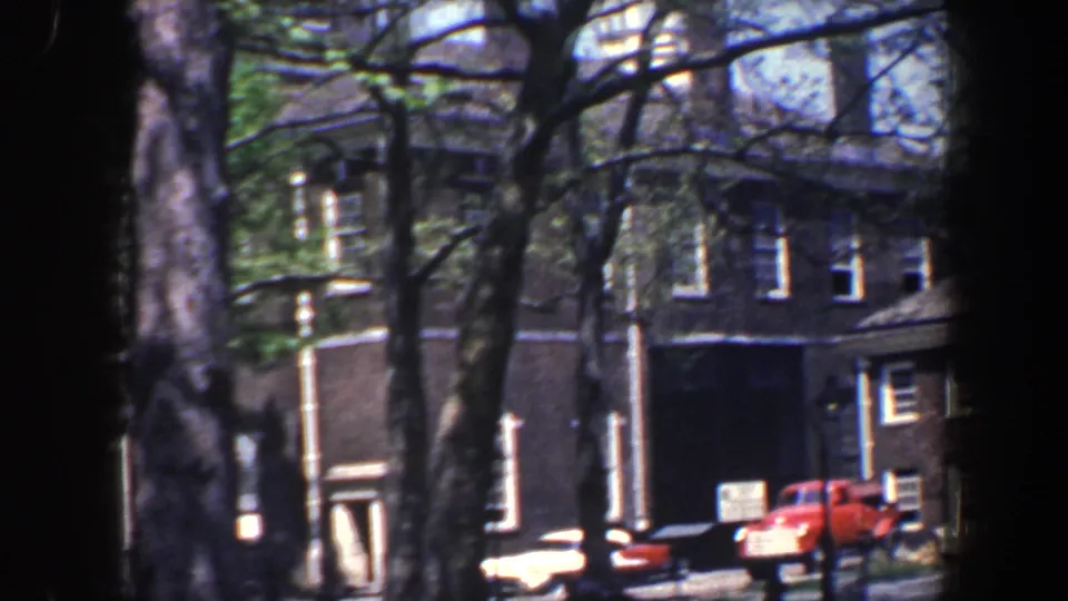 1962 Philadelphia street view with brick facades, parked cars, a red truck, and tree trunks partly blocking the 8mm frame.