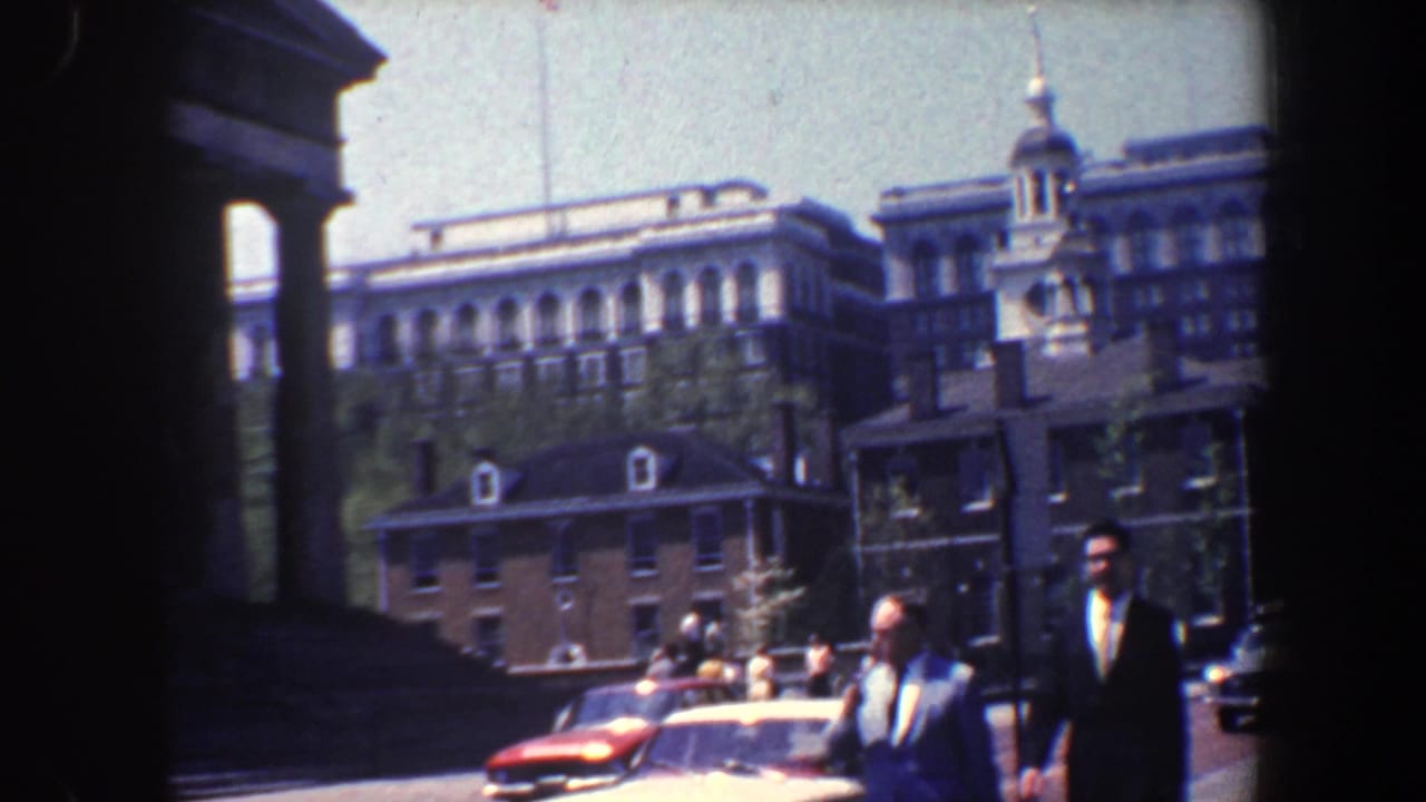 Men in suits walk near parked cars with historic buildings in the background.