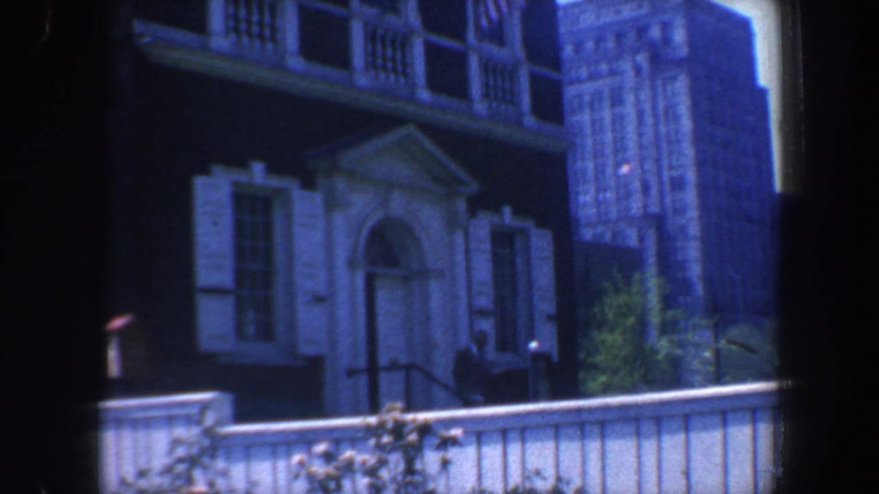 White fence and flowers below a dark brick building with arched doorway and tall city building behind.