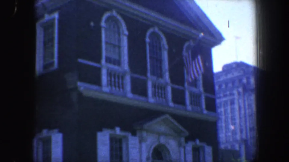 1962 Philadelphia home-movie frame looking upward at arched windows, white shutters, an American flag, and a downtown building beyond.