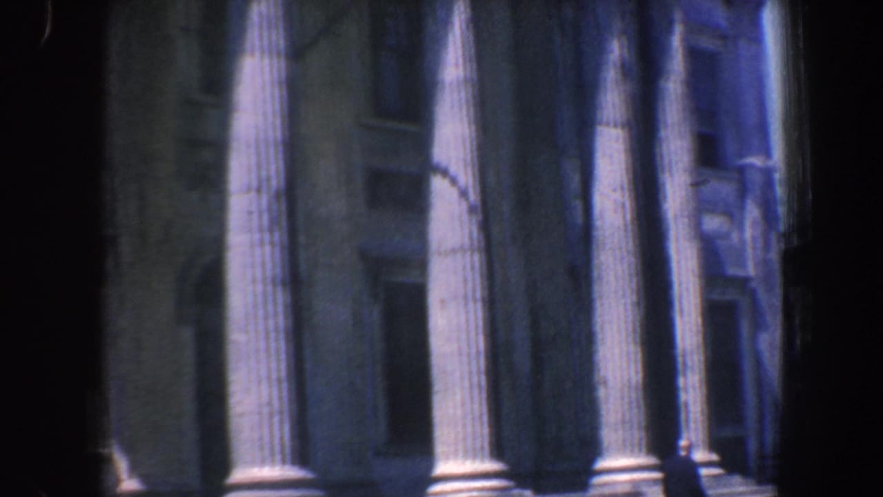 Columned facade with stone steps, windows, tree shadows, and a pedestrian near the lower right.