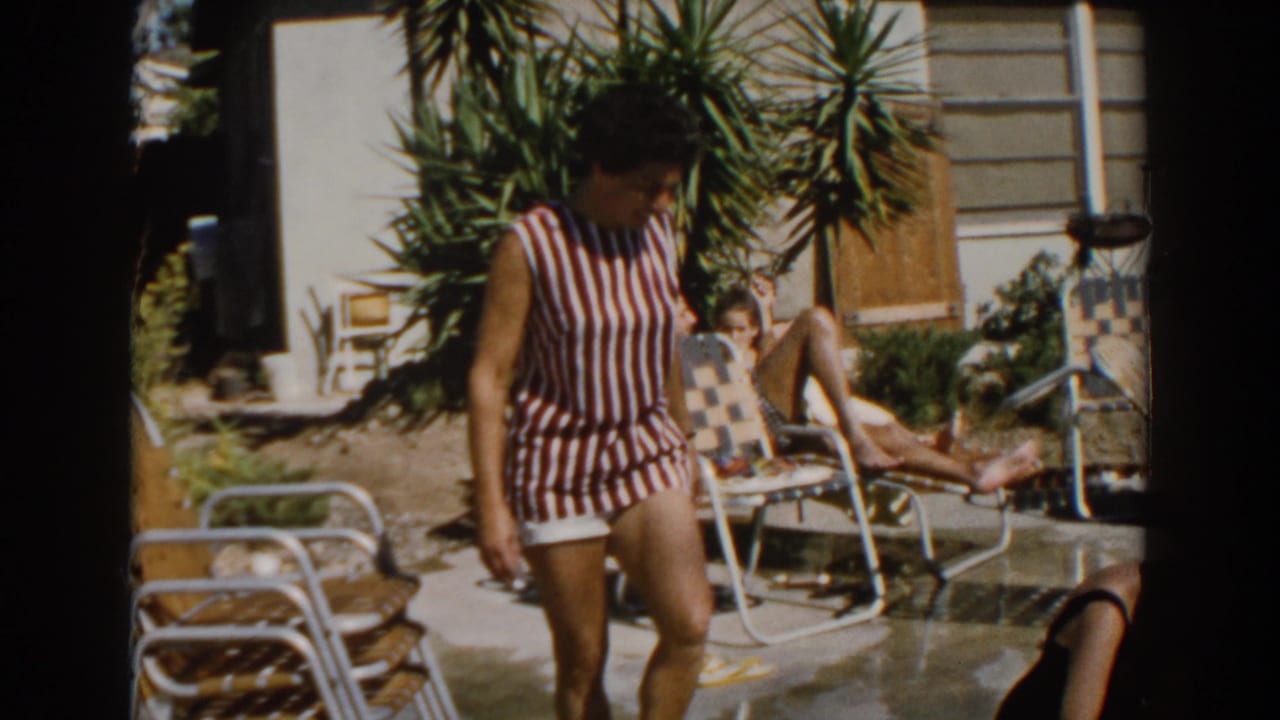 In North Hollywood in 1962, a woman in a striped sleeveless swimsuit walks past lounge chairs near a sunlit backyard pool.