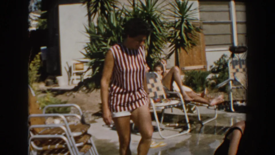 In North Hollywood in 1962, a woman in a striped sleeveless swimsuit walks past lounge chairs near a sunlit backyard pool.