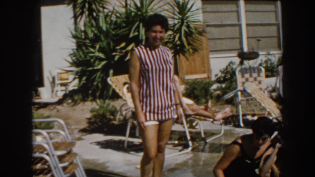 Woman in striped swimsuit standing near patio chairs and a backyard pool.