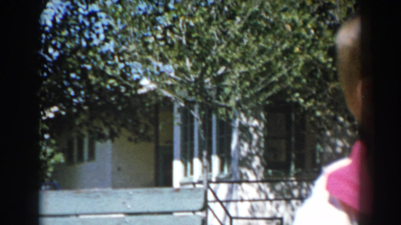 In a 1962 North Hollywood home-movie frame, a boy stands partly out of frame near a sunny lawn, hedges, and a light-colored house.