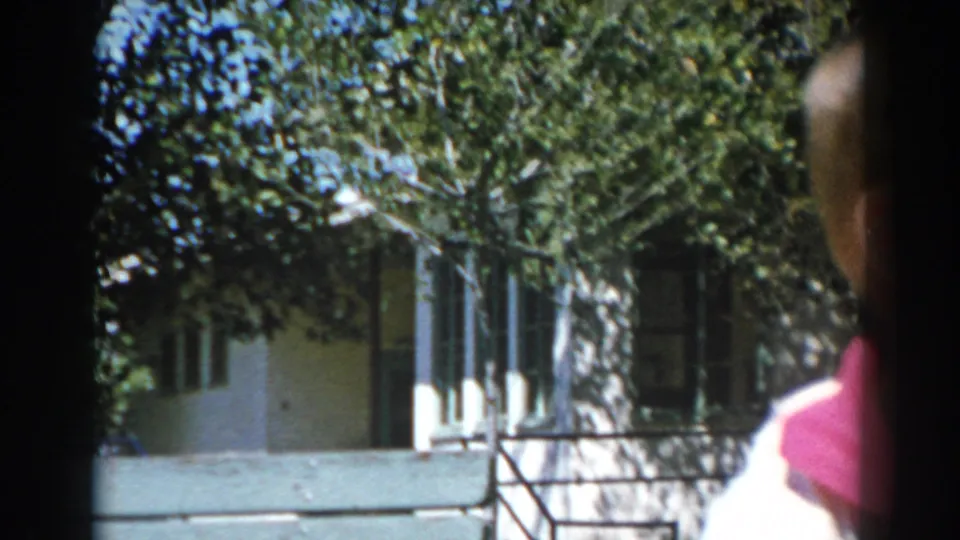 In a 1962 North Hollywood home-movie frame, a boy stands partly out of frame near a sunny lawn, hedges, and a light-colored house.