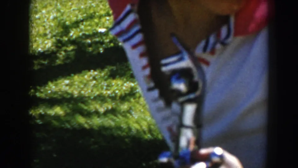 The boy leans into the frame over a small object, with striped shirt details, hands, grass, and hedge visible in the 8mm footage.