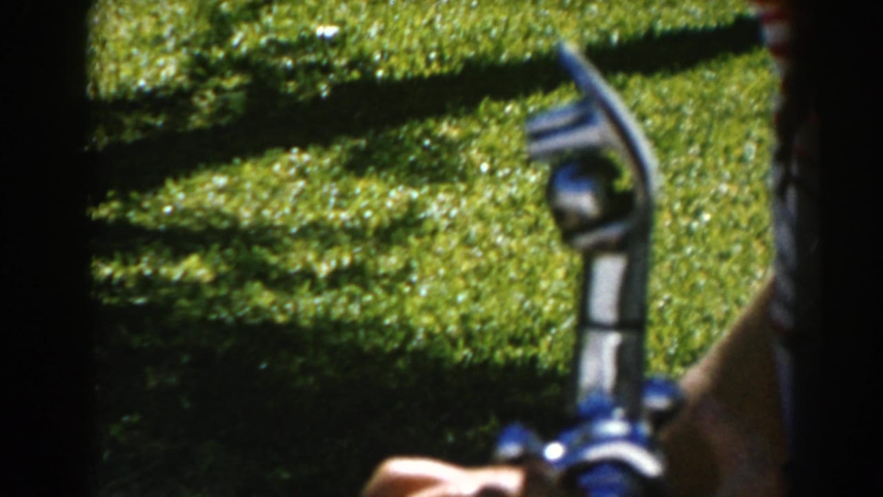 Hand holding a blue and silver drinking fountain nozzle above sunlit grass.