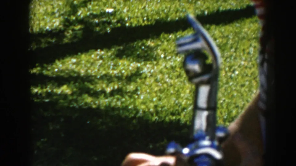 1962 North Hollywood 8mm frame showing a hand gripping a blue and silver drinking fountain nozzle against bright green grass.