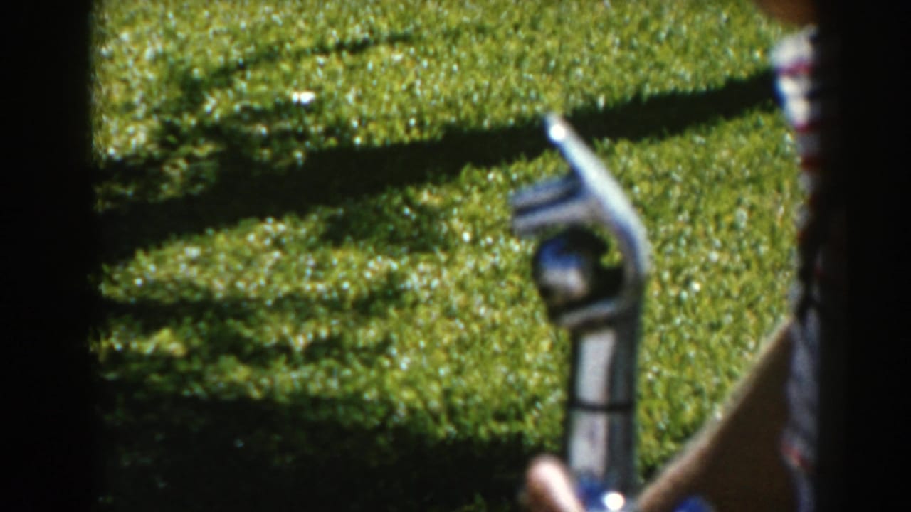 Drinking fountain nozzle held near grass, with a partial striped shirt at the edge.