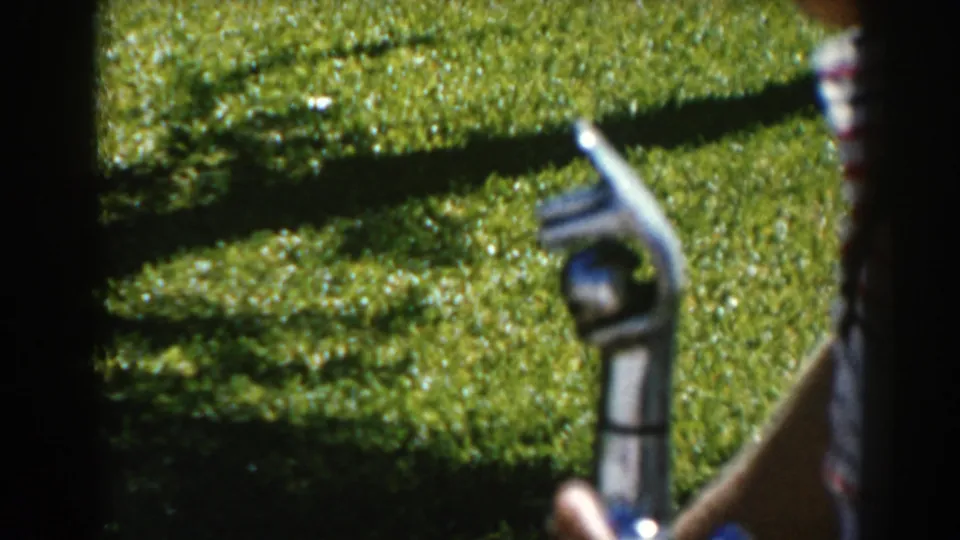 1962 North Hollywood home-movie frame with a hand holding a metal fountain nozzle near a white shirt with red stripes and sunlit grass.