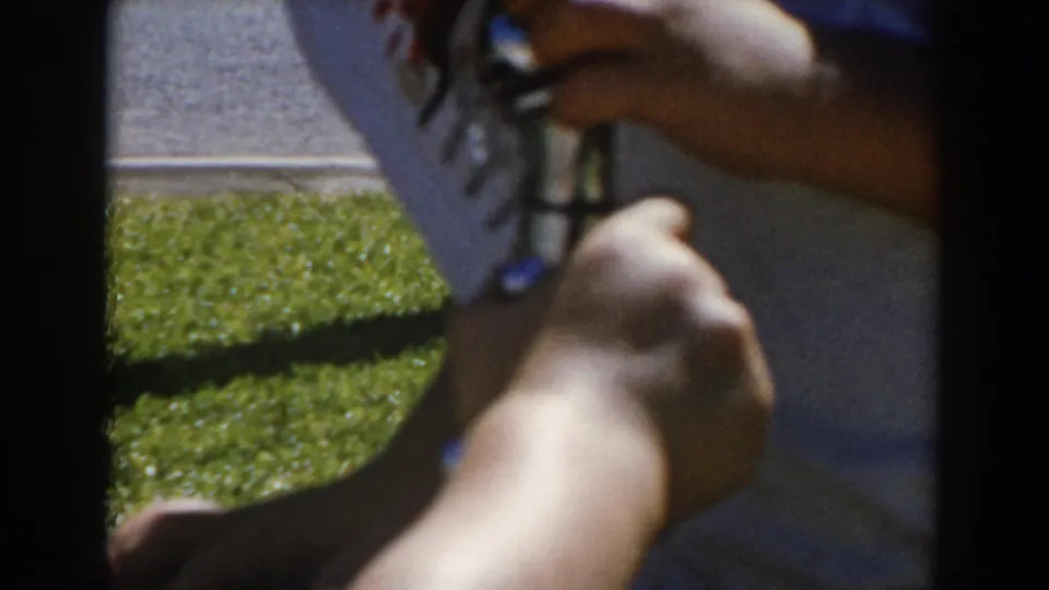 1962 North Hollywood 8mm frame showing a hand on the drinking fountain nozzle, with striped white shirt, grass, and pavement behind.