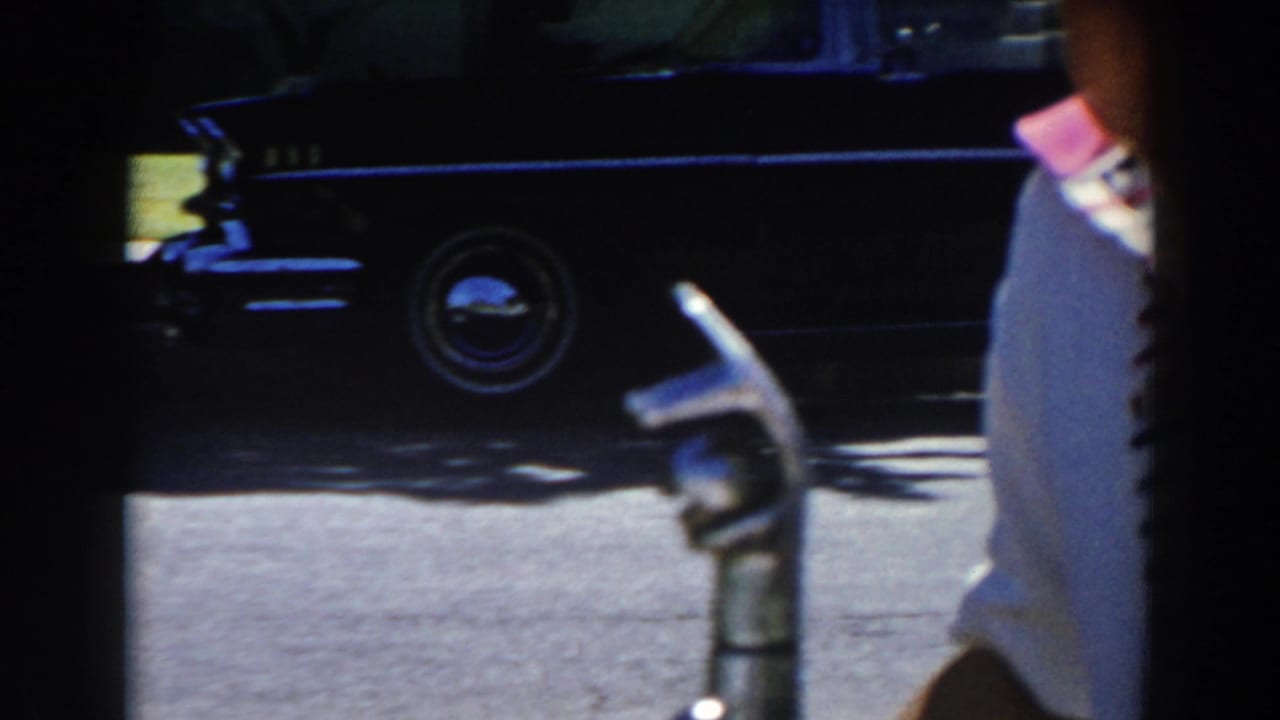 North Hollywood 1962 home-movie frame with a partial child in a striped shirt near a parked dark car and street curb.