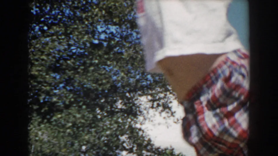 A child in a red plaid shirt and white shorts is partly framed against foliage in the 1962 North Hollywood home-movie shot.