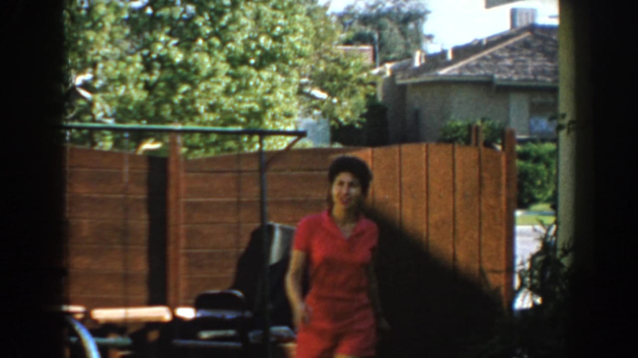 Woman in a red dress stands in a sunny backyard near a wood fence.
