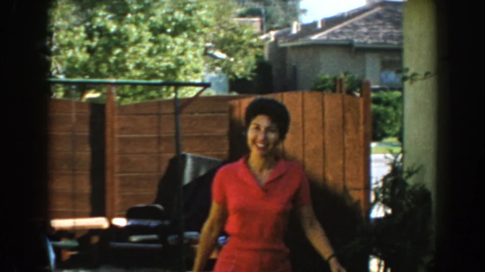A smiling woman in a red dress walks toward the camera in the North Hollywood yard, with a wood fence, trees and patio objects behind her.
