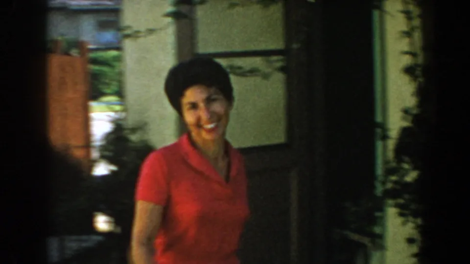 Close 8mm frame of the smiling woman in a red dress near the shaded side of a house, with fence and greenery visible behind her.