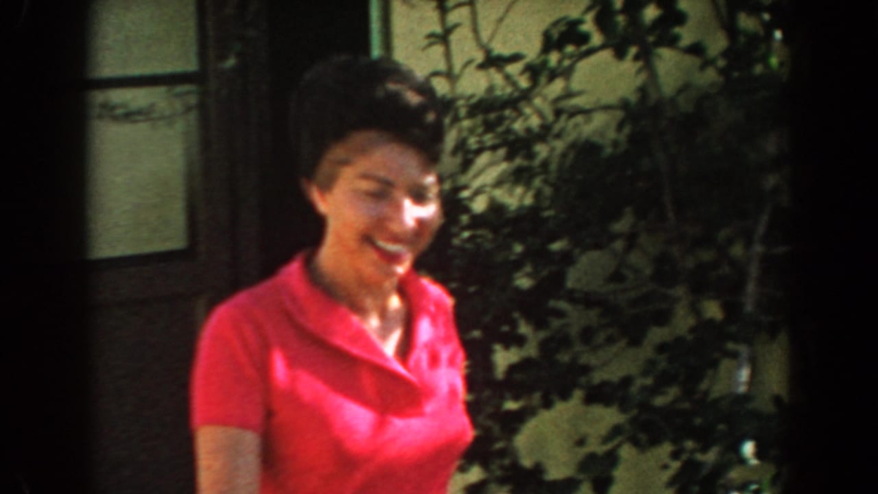 In 1962 North Hollywood home-movie footage, a smiling woman in pink walks past a doorway and leafy garden plants.