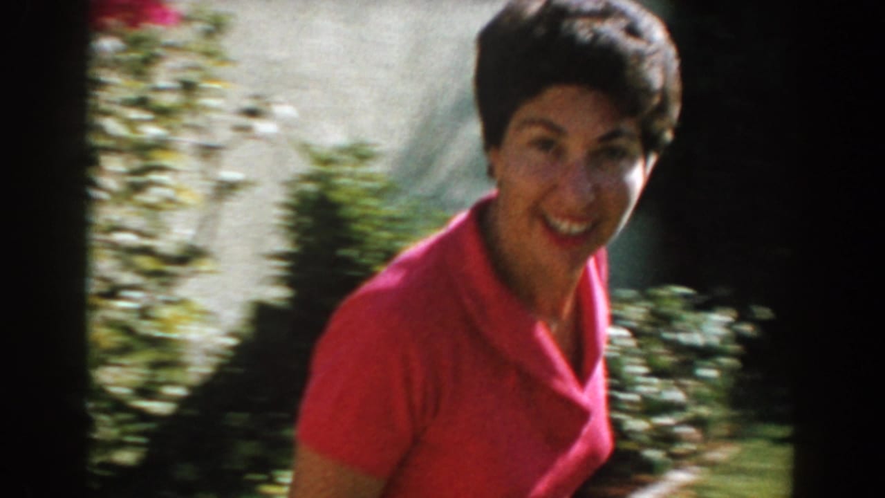Woman in a pink collared top smiles toward the camera near shrubs and a wall.