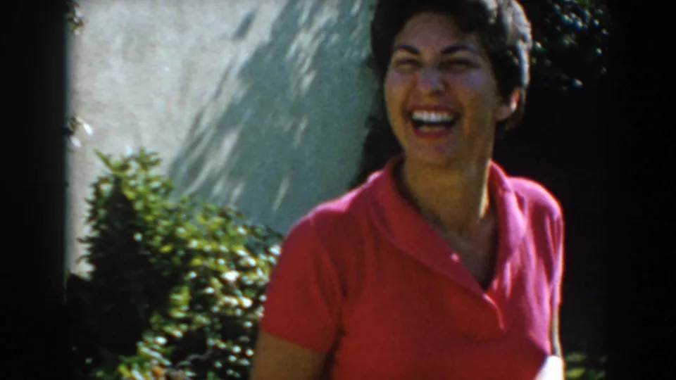 The woman smiles broadly in the garden, framed close against shrubs and a sunlit exterior wall.