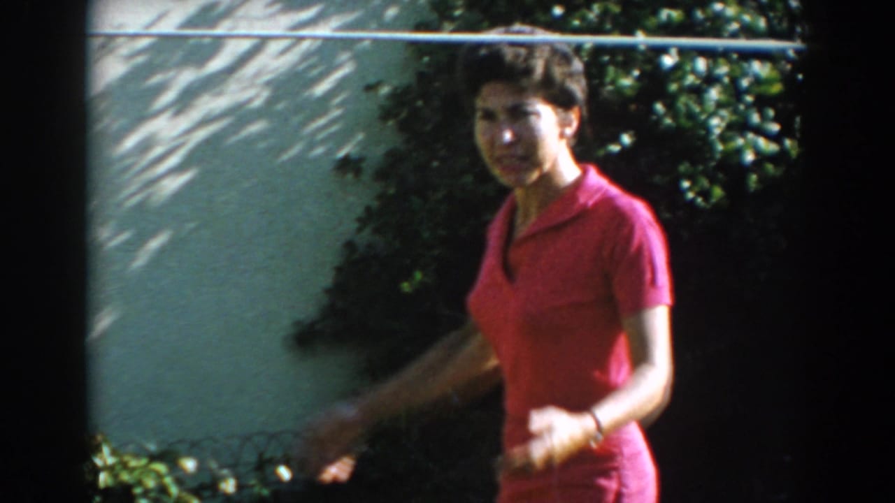 Woman in a red outfit walking near shrubs and a sunlit wall.