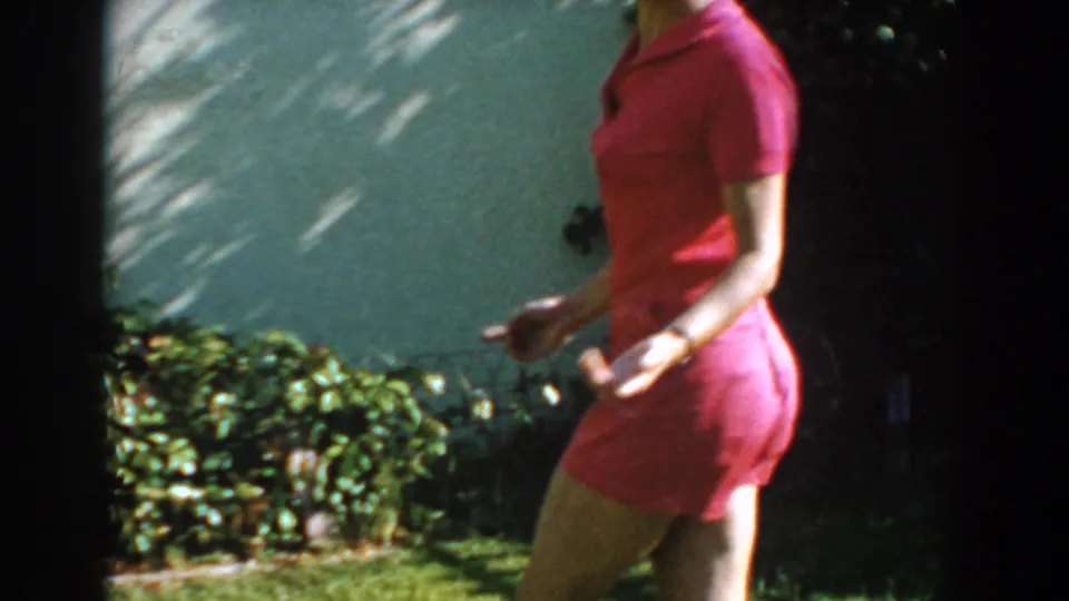 The woman in red moves through the right side of the 1962 backyard frame, with foliage, grass, and a pale exterior wall visible.