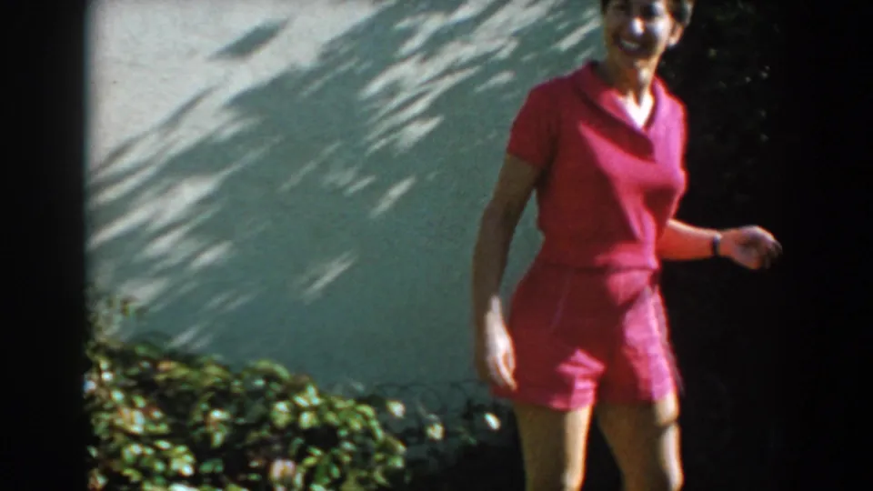 Adult woman in a red short-sleeve outfit walks toward the camera in the North Hollywood backyard, with wall shadows and low shrubs behind her.