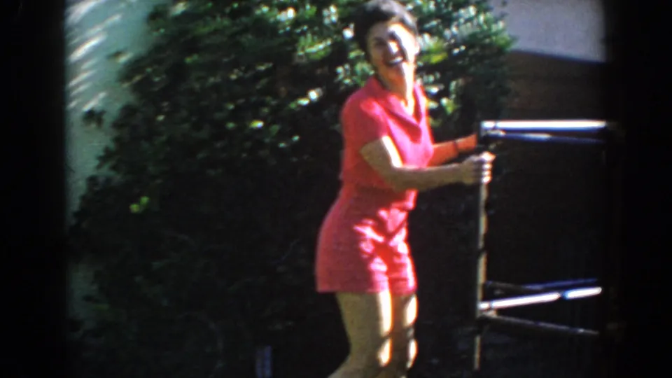 Smiling woman in a red outfit stands near a dark outdoor chair or patio frame in a sunny 1962 North Hollywood backyard.