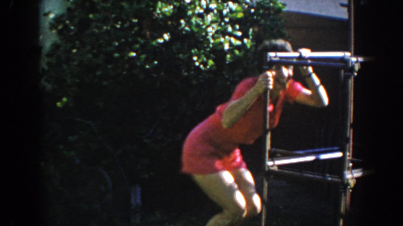 Woman in a pink short suit crouches beside a metal outdoor frame in a sunny yard.