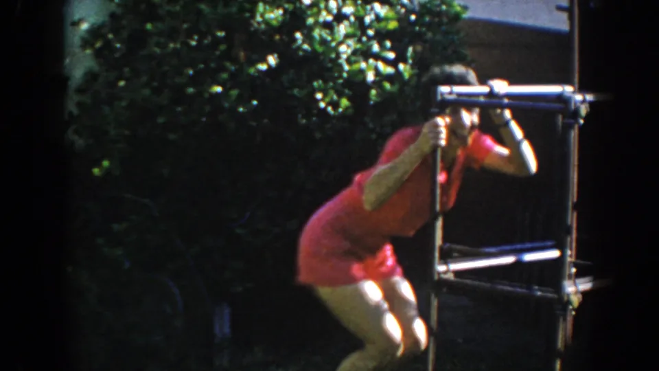 In 1962 North Hollywood home-movie footage, a woman in a pink short suit crouches playfully near a metal frame in a sunlit yard.